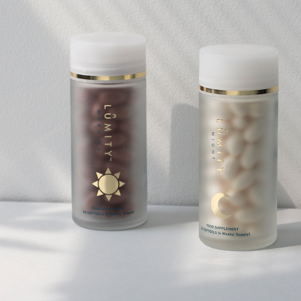 Lumity Morning and Night frosted supplement bottles with white caps and gold trim; left dark brown softgels, right white.