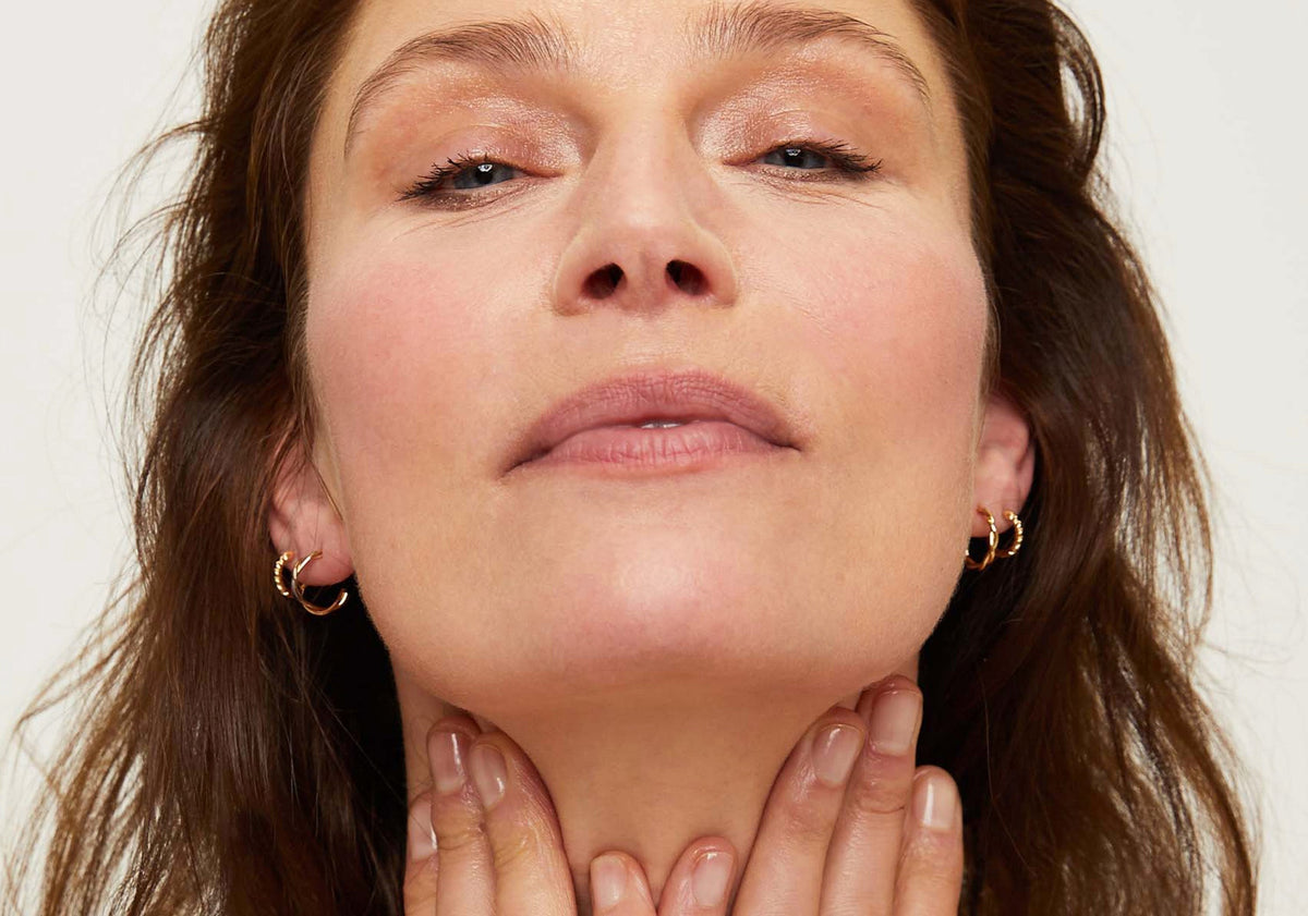 Close-up low-angle view of small twisted-design gold hoop earrings worn by a woman, hands at her neck