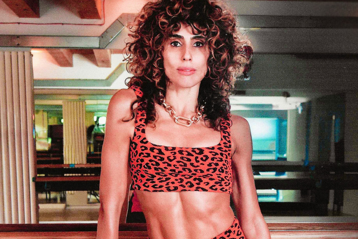 Adult woman in red leopard-print sports bra and matching leggings, gold chain, standing before mirrored gym wall.