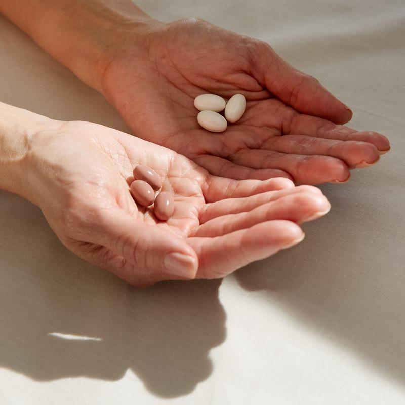 Close-up of two open hands holding three beige capsules in left palm and three white capsules in right palm.