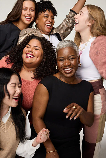 Five diverse adult women standing close together, smiling and laughing against a plain white background, mid-torso framing.