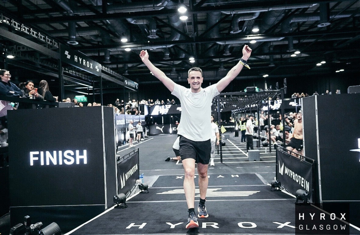 Male runner in white t-shirt and black shorts crossing HYROX finish line with arms raised