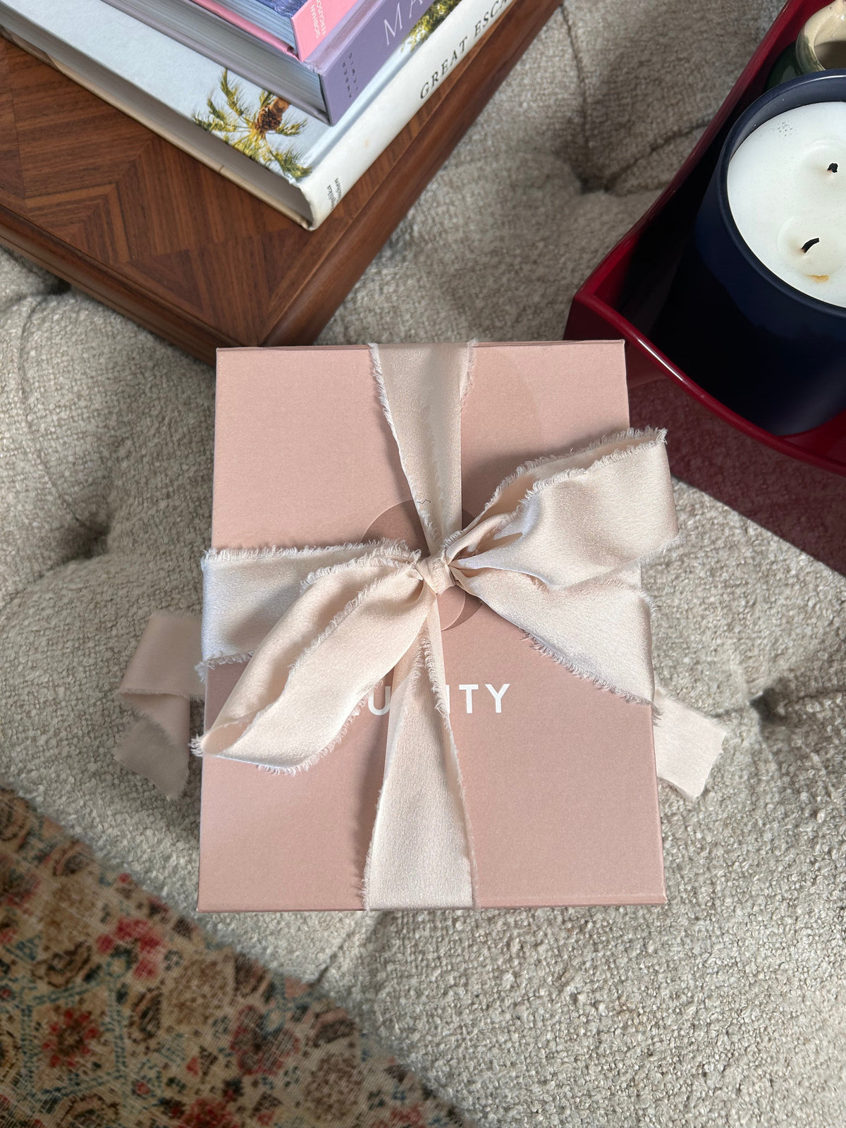 Light pink gift box with off-white frayed-edge ribbon bow on a tufted beige surface, angled top-down view.