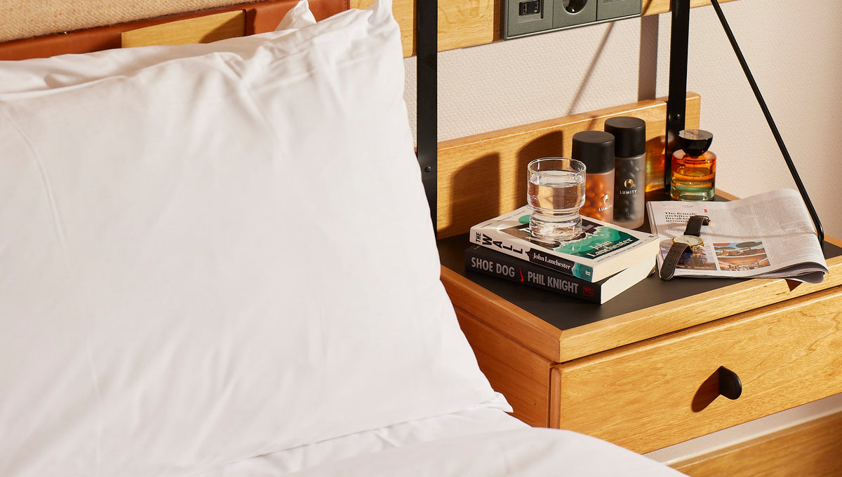 Two cylindrical Lumity bottles with black caps on a wooden bedside table beside books and a glass of water