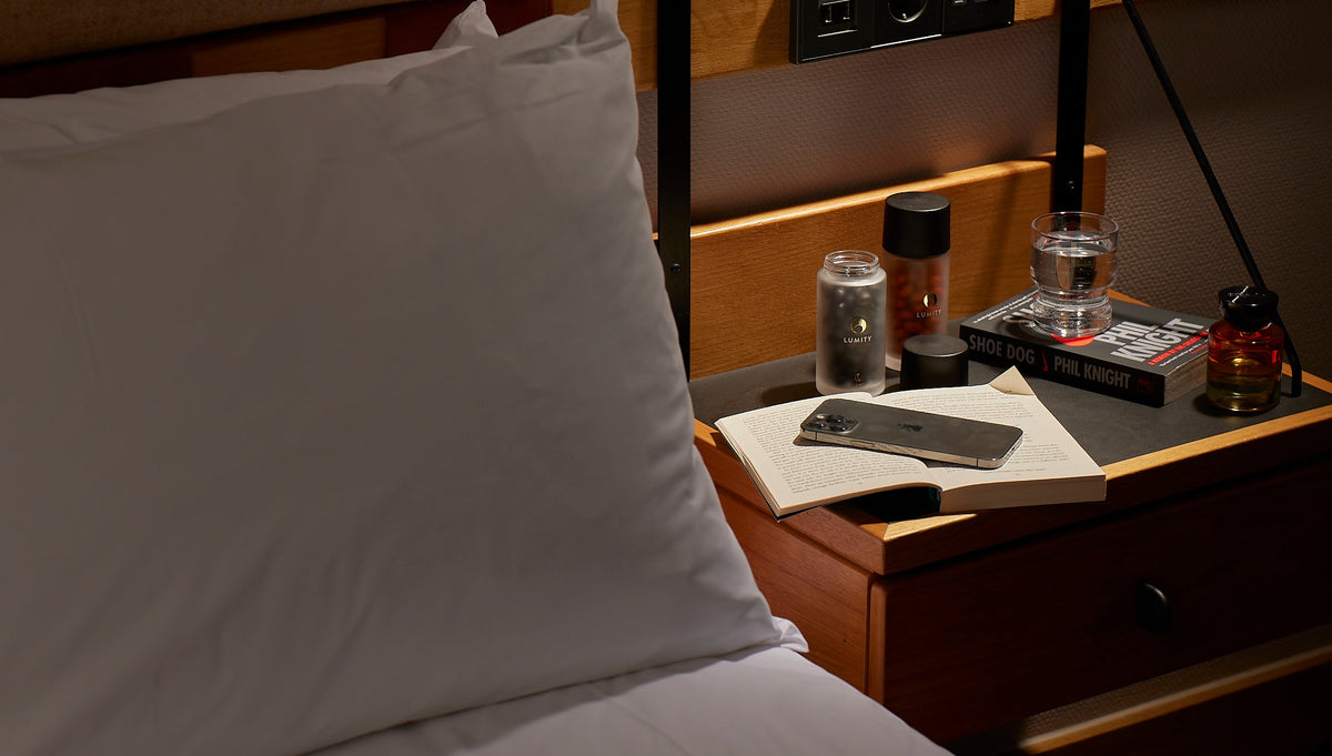 Two frosted Lumity bottles with black caps and an amber dropper bottle on a wooden bedside table by a white pillow