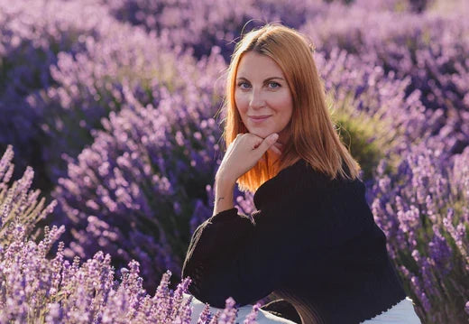 Adult woman with reddish hair seated among purple lavender, wearing a black long-sleeve top and white pants, hand under chin.