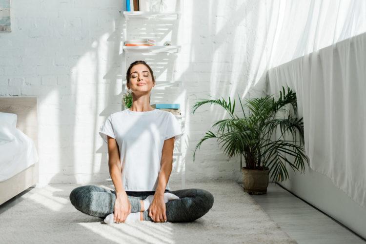 Young woman meditating cross-legged on light carpet, eyes closed, wearing white T-shirt and gray patterned leggings.
