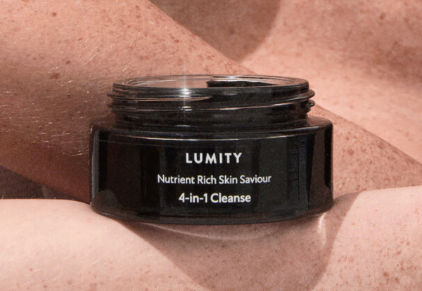 Black round Lumity jar labeled 'Nutrient Rich Skin Saviour 4-in-1 Cleanse' resting centered on a freckled forearm, close-up
