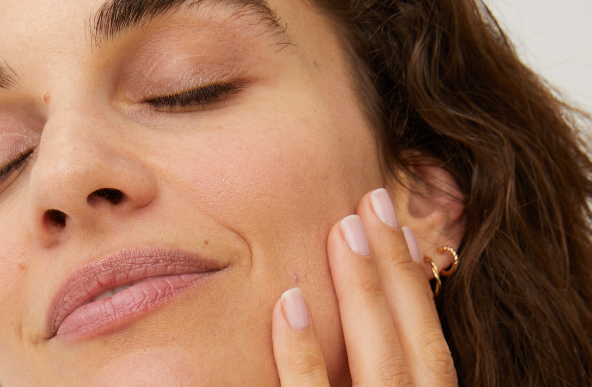 Close-up of a woman's right-side face with eyes closed, hand gently on cheek, natural skin, pink nails, gold earrings.
