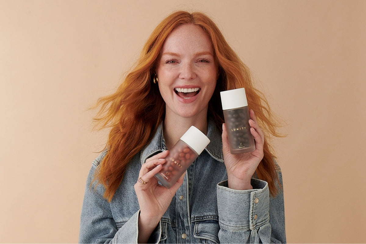 Smiling woman in a light denim jacket holding two frosted LUMITY supplement bottles with white caps near her face