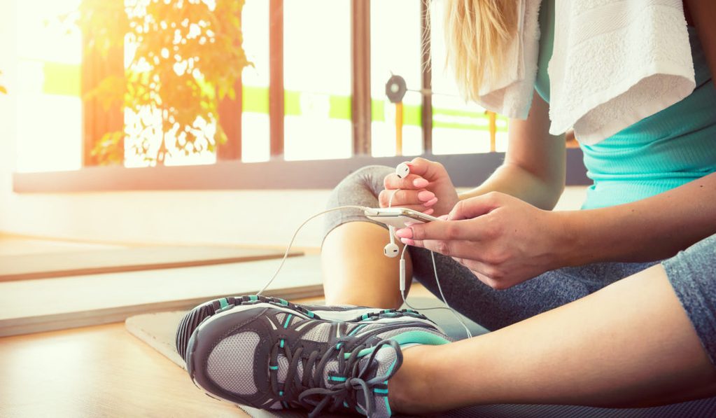 Young woman seated on a gym floor in a turquoise sleeveless top and gray leggings, holding a smartphone and white earphones.