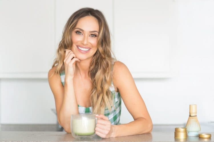 Woman in a green-and-white checkered sleeveless top smiles while holding a glass mug of green beverage at a white counter