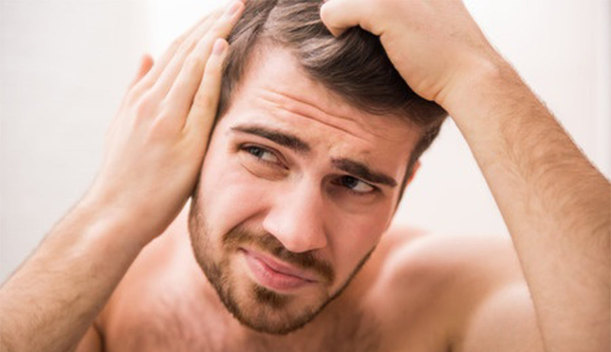 Shirtless young man with short brown hair and beard touching his scalp, pensive close-up with soft lighting.