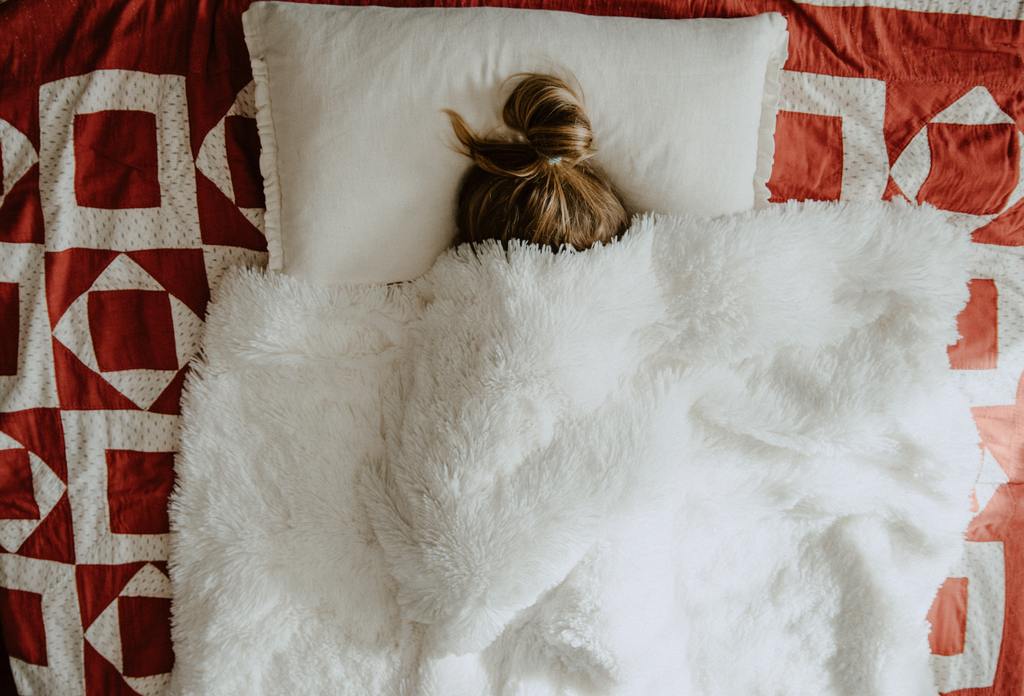 Overhead view of a person tucked under a fluffy white blanket, topknot visible, red-and-white geometric bedspread.
