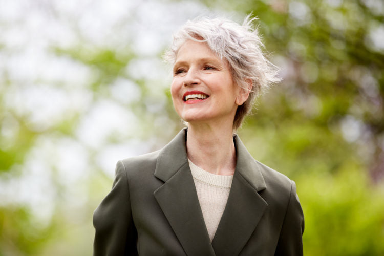 Older woman with gray hair in an olive blazer and cream sweater, smiling and looking right against blurred green foliage.