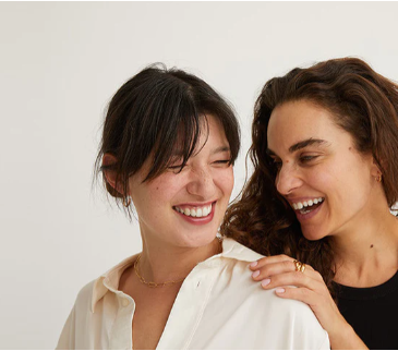 Two women close together, left in a white button-up smiling with eyes closed, right laughing with hand on her shoulder.