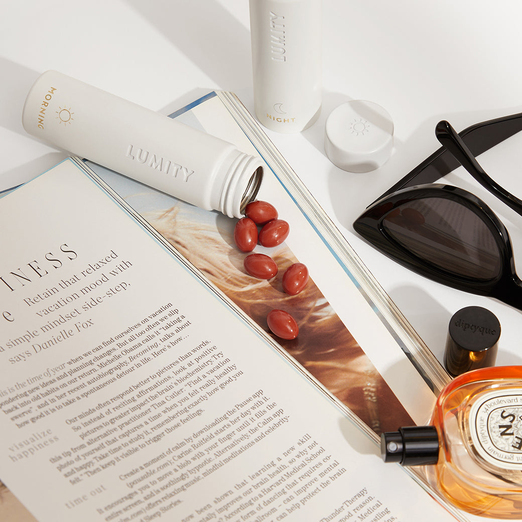 Red capsules spilling from a white Lumity Morning bottle beside sunglasses, perfume, and an open wellness magazine.