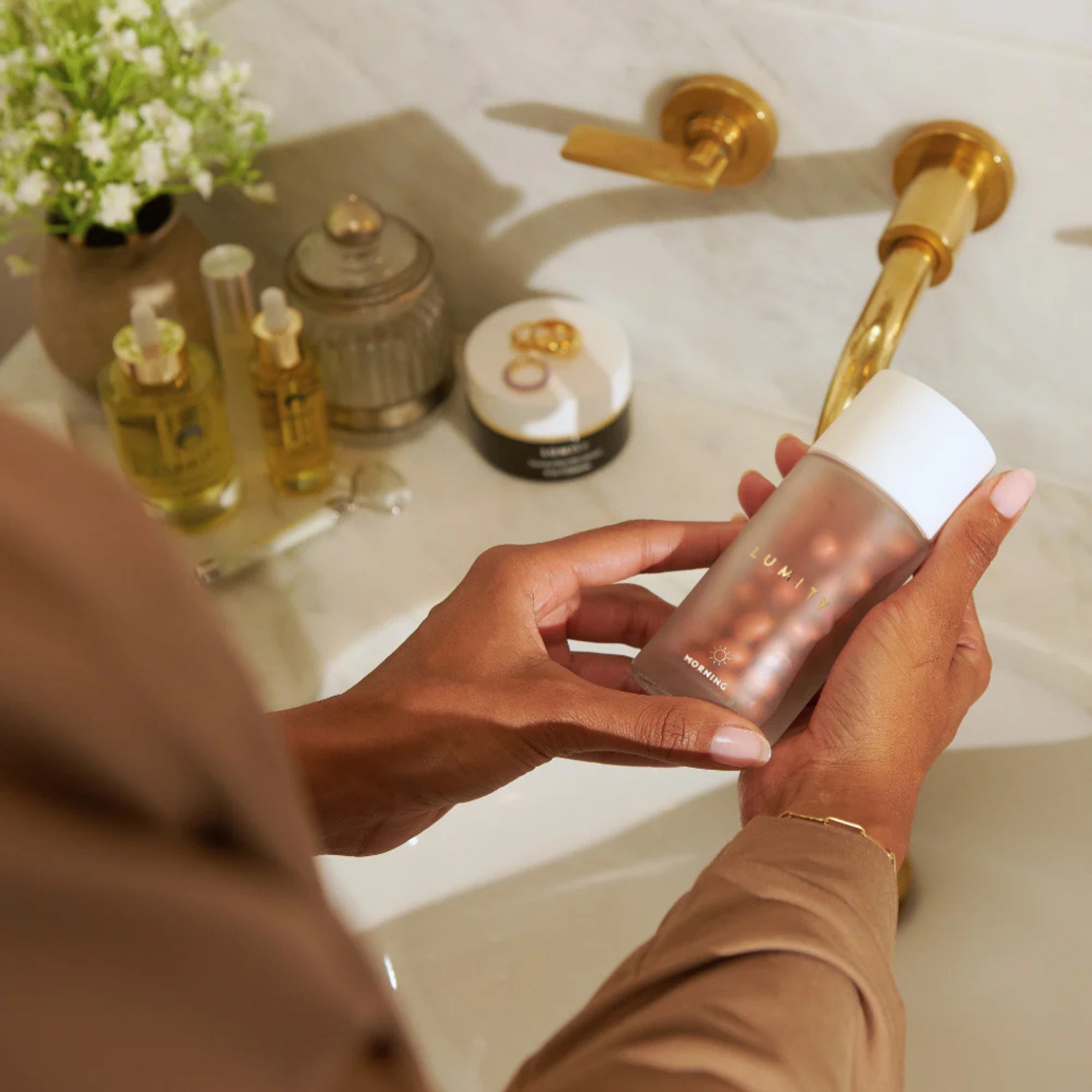 Hands holding a frosted bottle of Lumity Morning & Night Women's Supplement over a bathroom sink with wellness products nearby