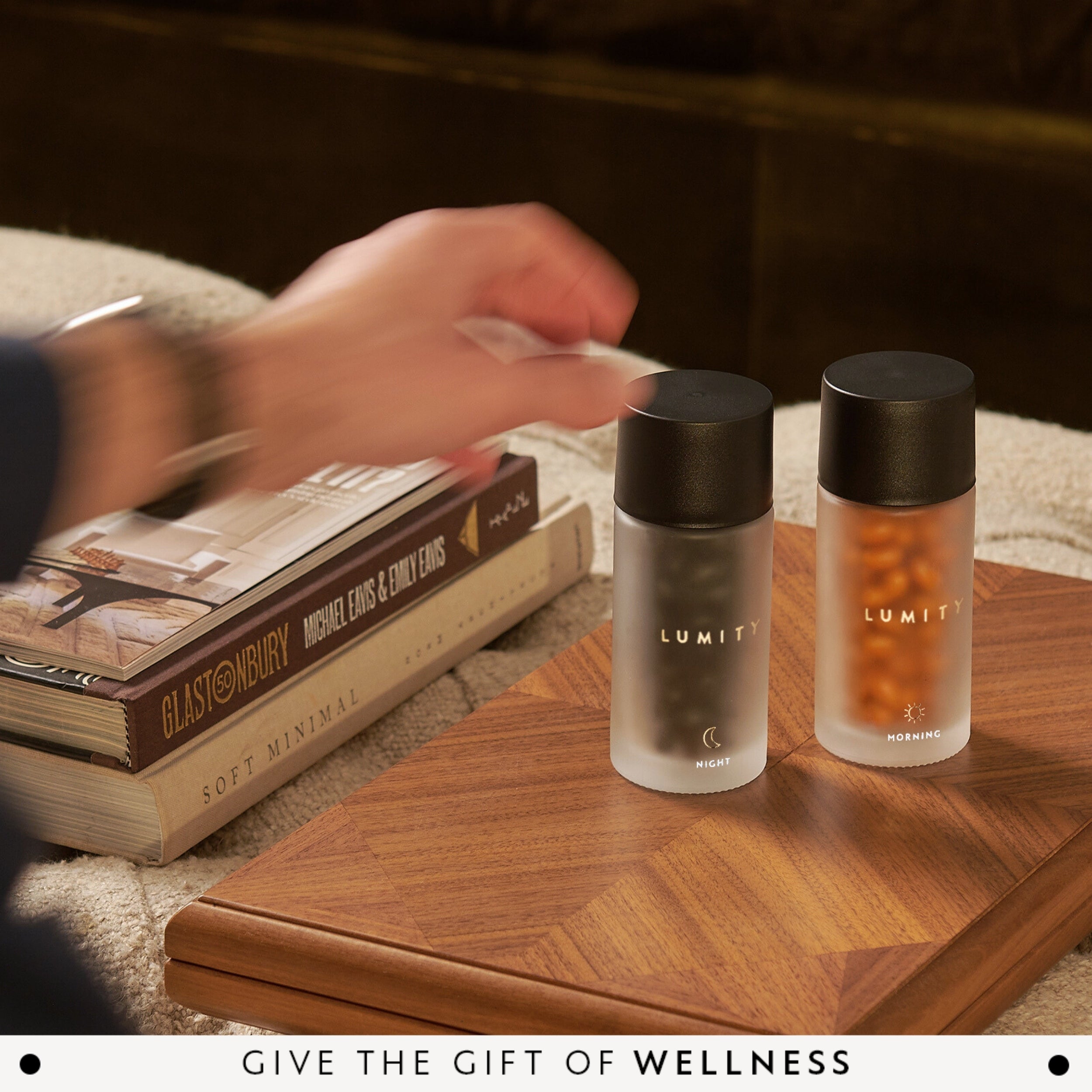 Morning and night Lumity supplements in frosted and amber glass bottles placed on wooden tray by books and hand reaching to open bottle
