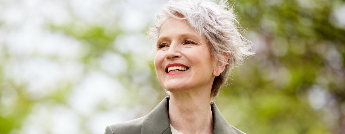 Older woman with short white hair smiling and looking left, wearing a muted green blazer in natural light.