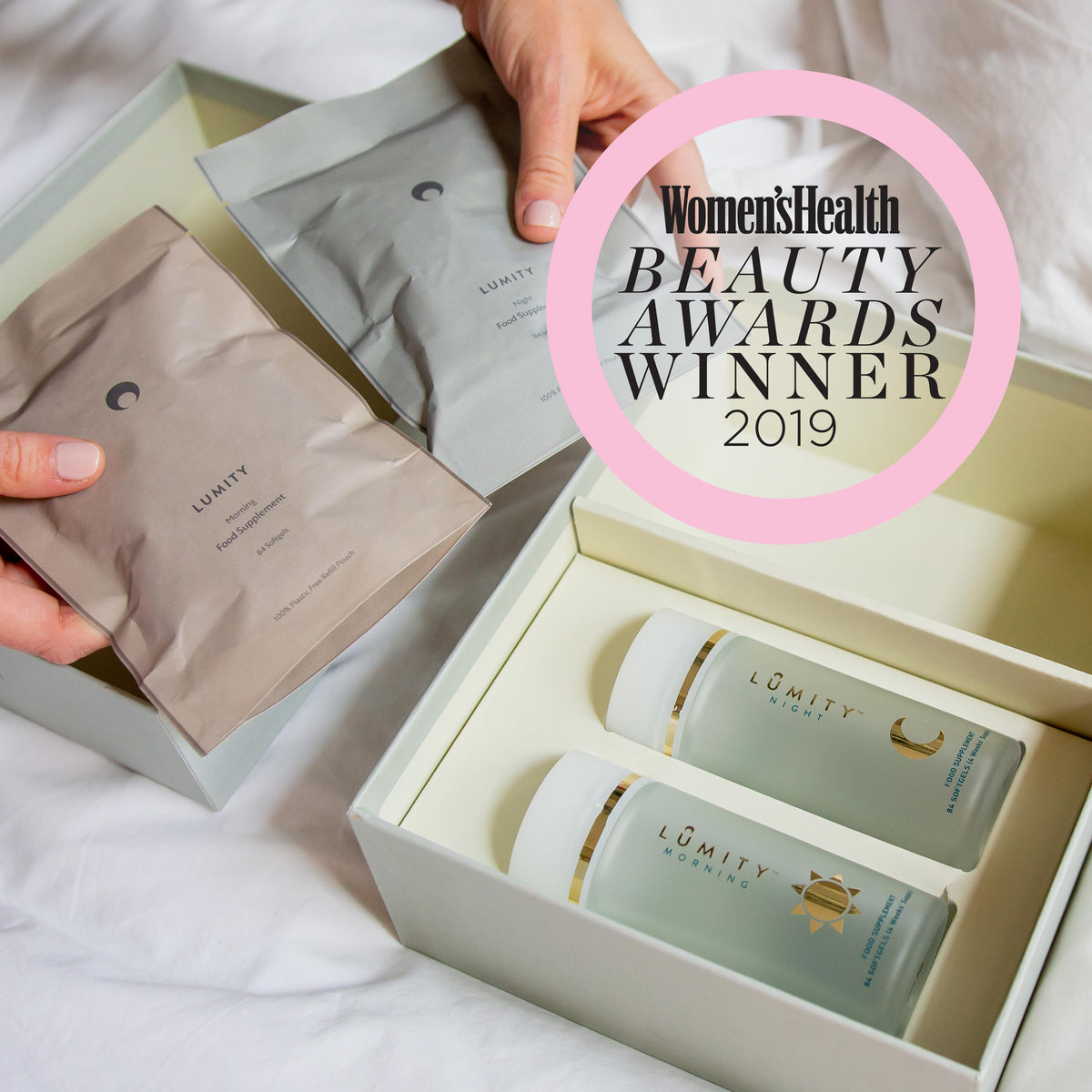Top-down close-up: hands hold beige & gray Lumity Morning & Night pouches over an open box with frosted glass bottles.