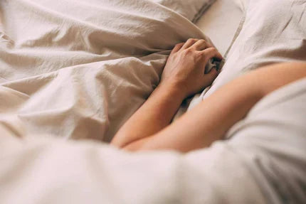Hands resting on beige bed sheets, fingertips gripping soft, wrinkled fabric in warm natural light.