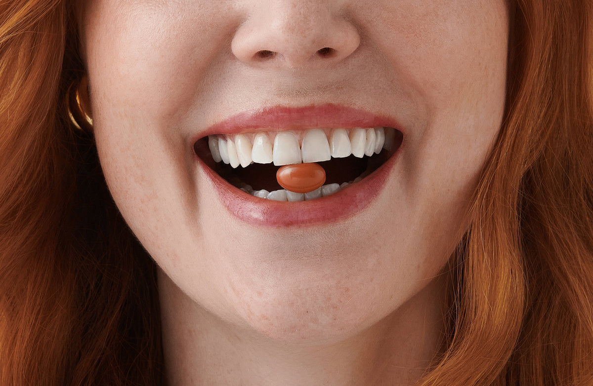 Smiling red-haired woman holding an oval orange pill between her teeth, close-up on mouth and freckled skin.