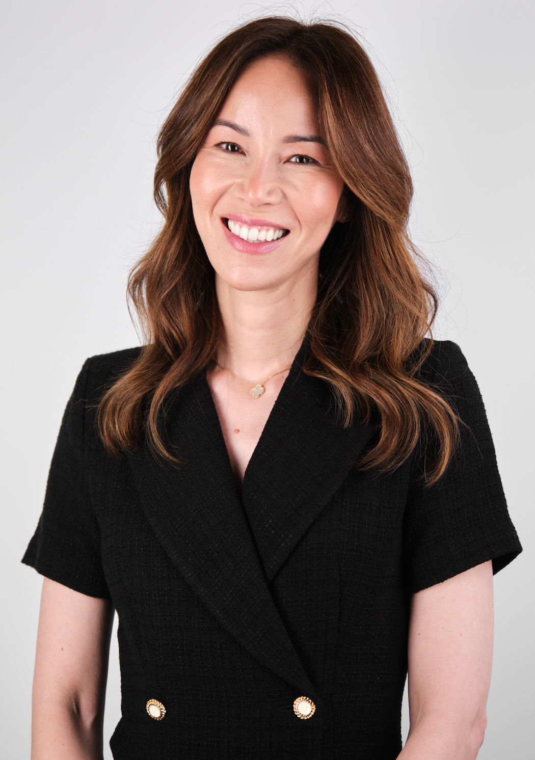 Woman wearing black textured top with gold buttons and a gold clover pendant necklace, smiling.