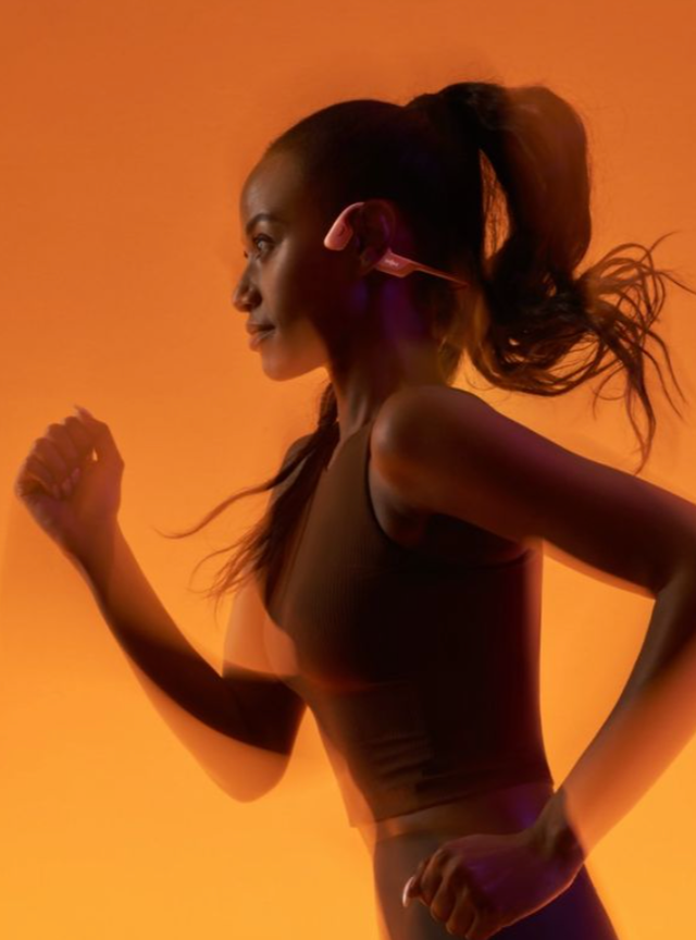 Side profile of a dark-skinned woman jogging in a sleeveless top, ponytail trailing, earbuds against warm orange background