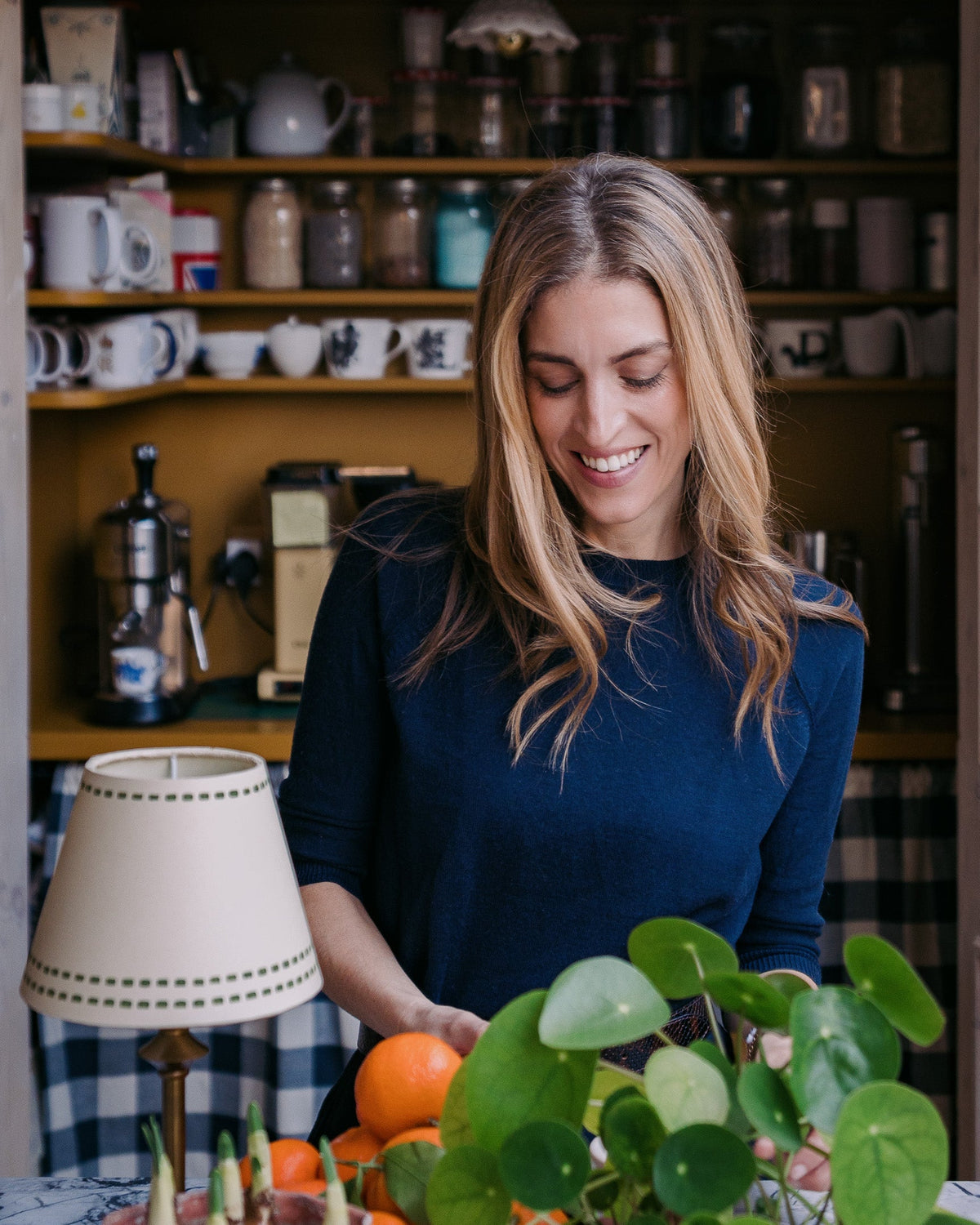 Woman in a dark blue long-sleeve top smiling and looking down at oranges and green plants on a kitchen table.