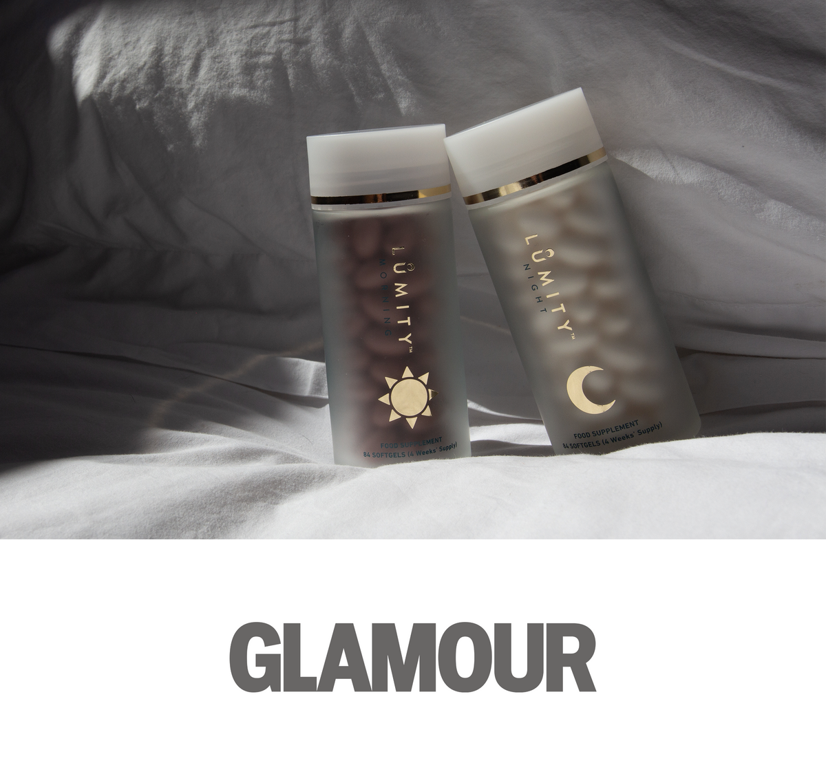 Two frosted Lumity supplement bottles, Morning (gold sun) and Night (gold moon), showing softgels, leaning on white fabric.