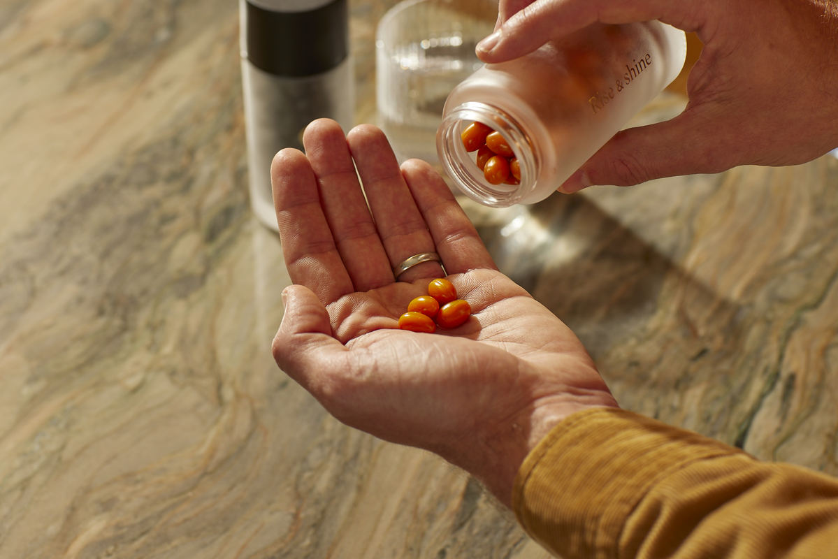 Hands at eye-level pouring small orange capsules from a glass 'Rise & shine' jar into an open palm, yellow sleeve visible
