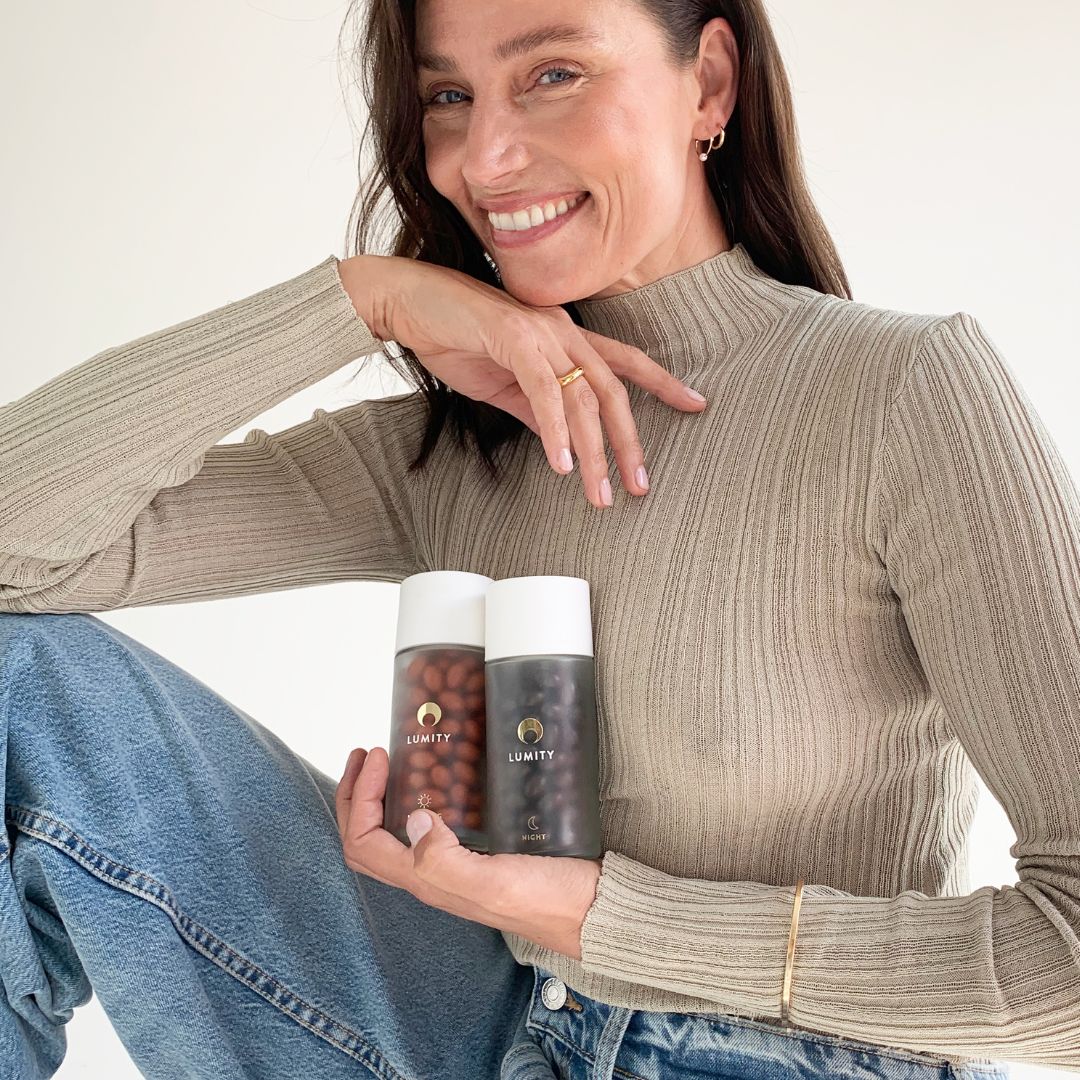 Lumity supplement bottles held to chest by smiling woman in beige ribbed turtleneck, white caps and brown and darker capsules