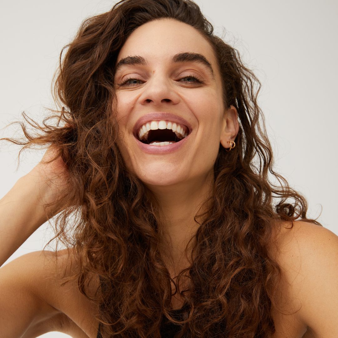 Young woman with long curly brown hair smiling broadly, wearing small gold hoop earrings and a sleeveless black top.