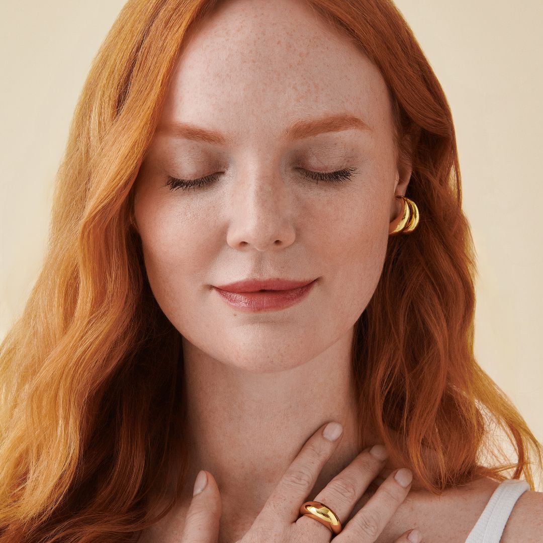 Woman with long red hair and freckles, eyes closed, wearing white top and gold hoop earrings, hand resting on her chest.