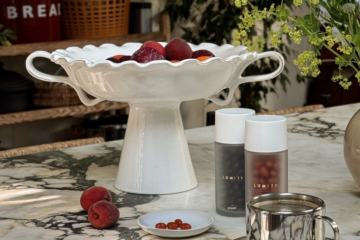 Frosted Lumity bottles (one marked NIGHT) beside a white pedestal bowl of red-orange fruit and a dish of capsules.