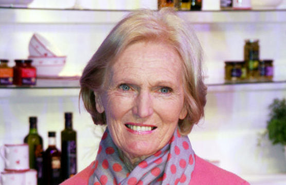 Older woman with blonde hair and blue eyes in a pink top and gray scarf with pink polka dots, kitchen shelves behind.