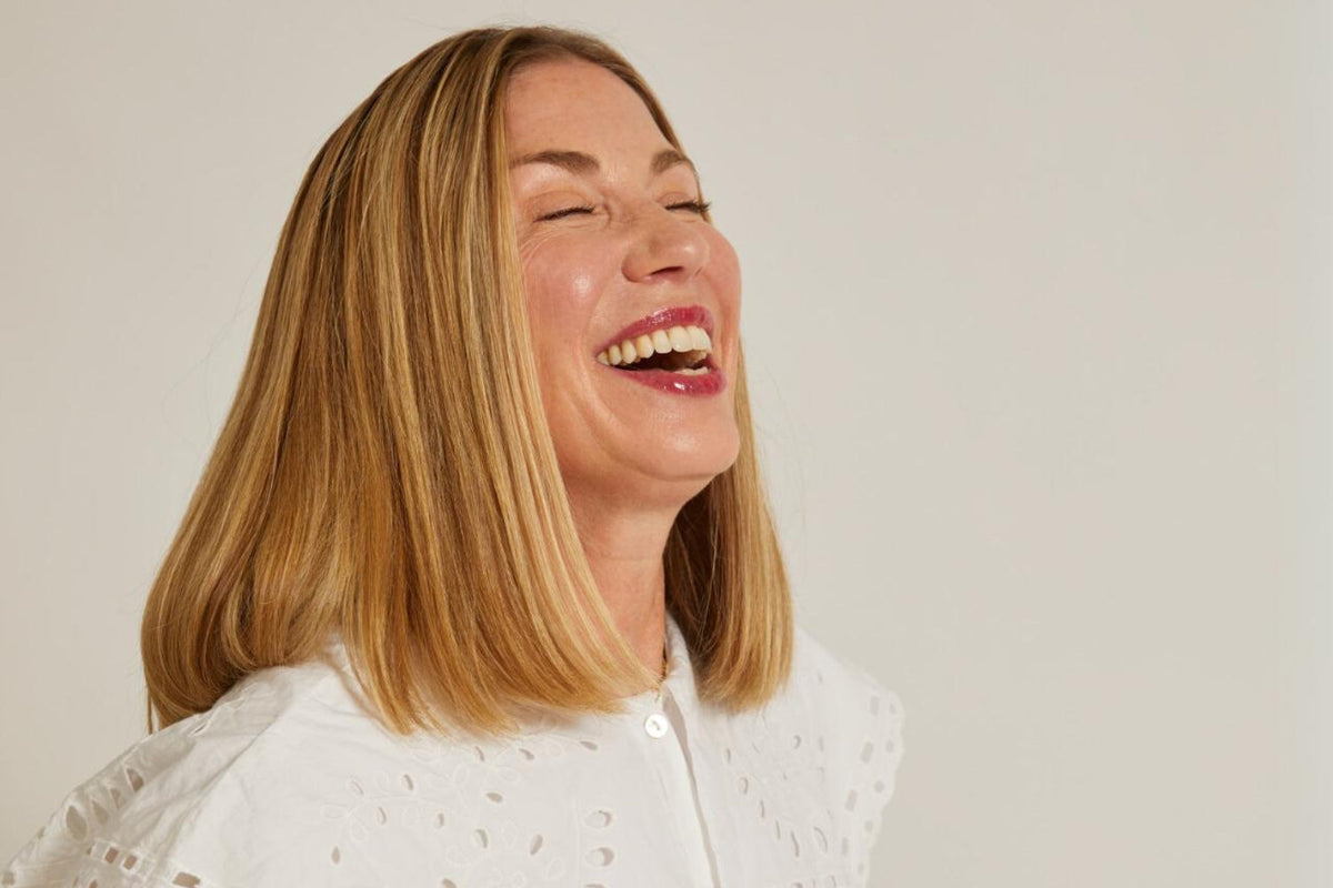 Close side profile of a middle-aged blonde woman laughing with eyes closed, wearing a white eyelet-collared blouse.