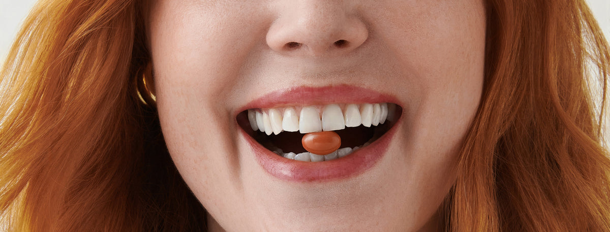 Close-up of a smiling woman with red hair showing an orange oval pill resting on her tongue.