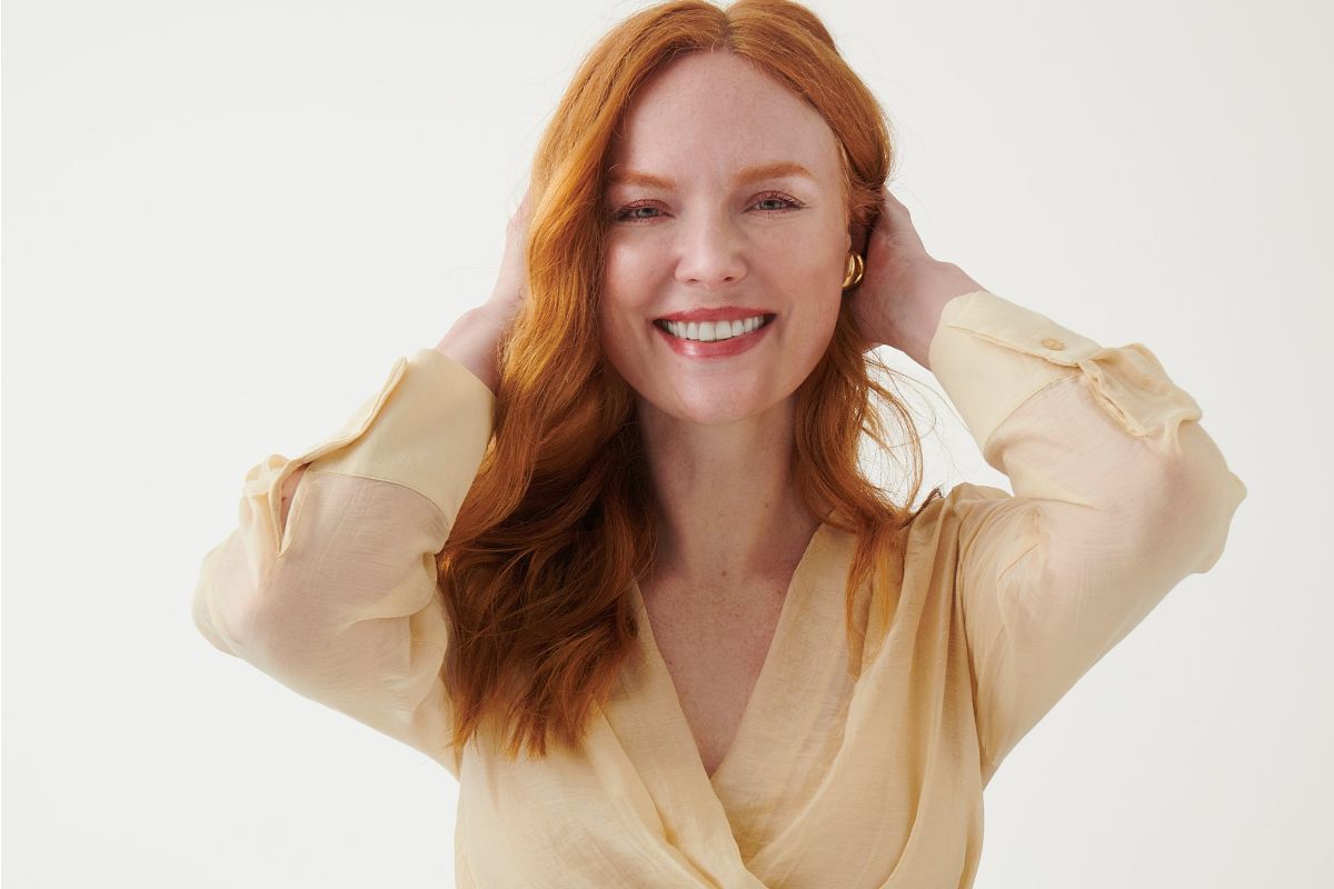 Woman wearing a light beige semi-sheer long-sleeve blouse, close-up mid-shot, smiling with hands behind her head.