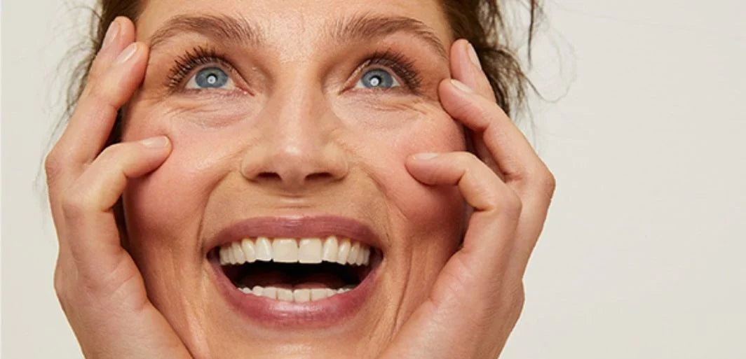 Light-skinned woman with blue eyes smiling, hands gently cupping her cheeks in a close-up portrait.