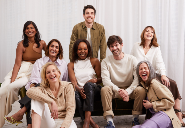 Seven diverse adults smiling in two rows on and around a dark green velvet sofa against a white curtain backdrop.