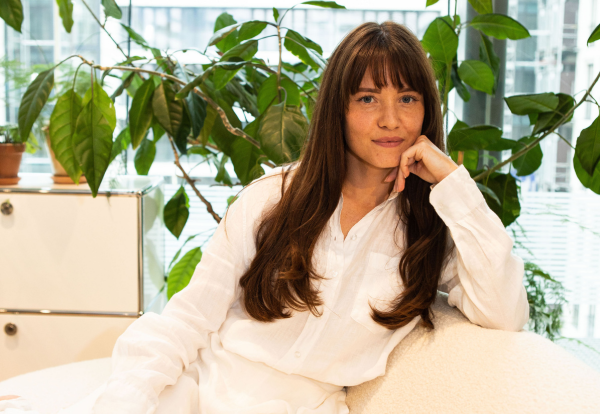 Young woman in a white button-up blouse seated on a light textured chair, resting her left hand on her cheek.