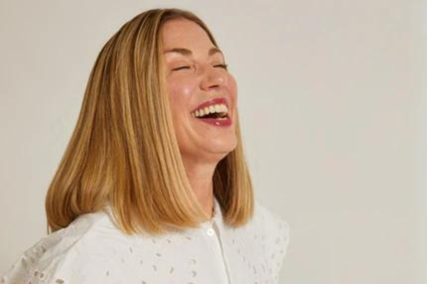 Laughing light-skinned woman in a white eyelet blouse, shoulder-length blonde hair, eyes closed, close-up portrait.