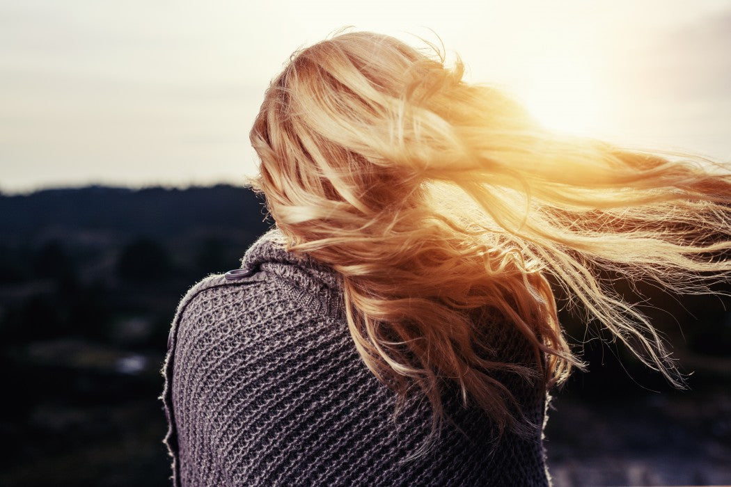 Back view of a woman in a chunky gray knit sweater, long blonde hair blown right, sunlit blurred hills behind.