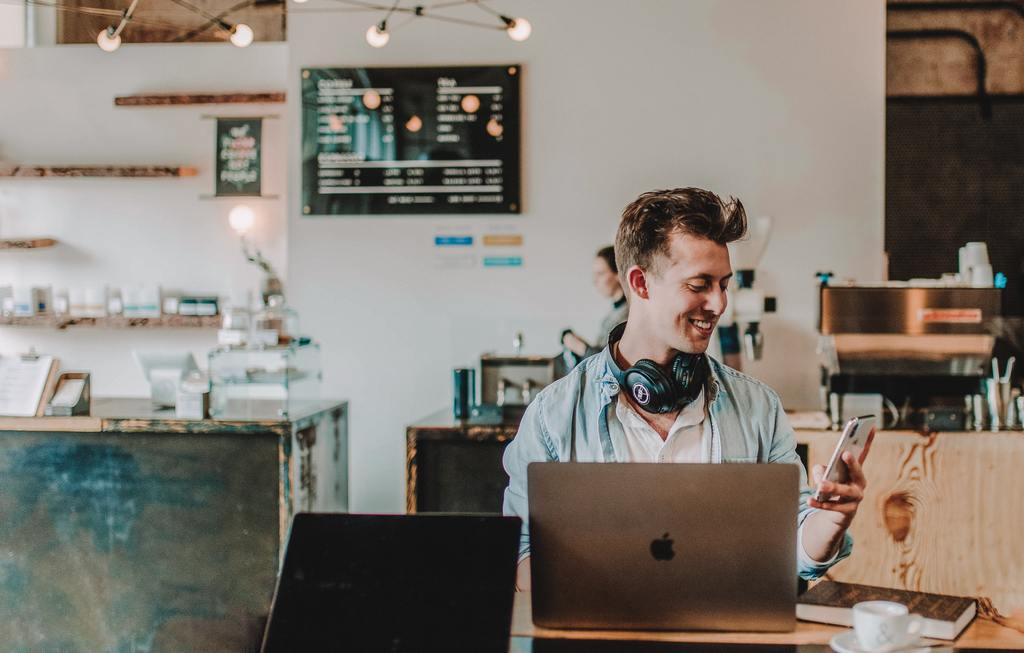 Young man in light denim shirt with headphones, smiling at his smartphone next to a silver laptop and coffee cup