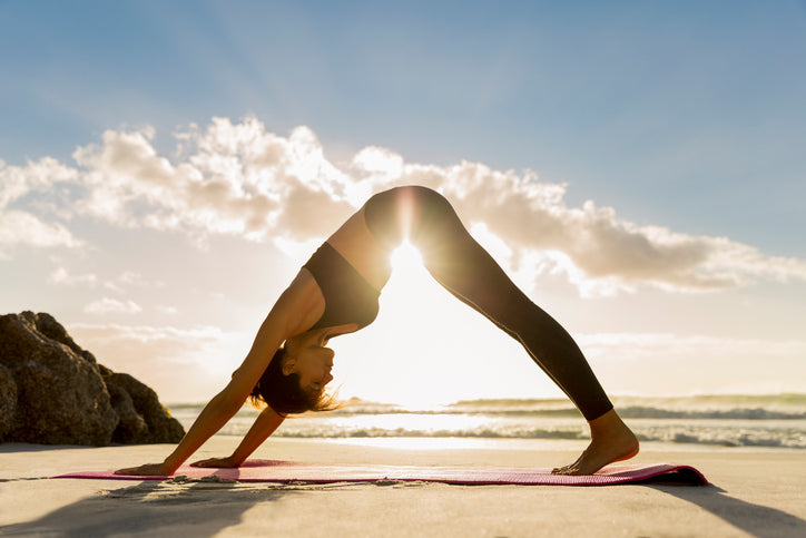 Pink yoga mat on sandy beach with woman in black holding downward dog at sunrise, sun backlighting her silhouette