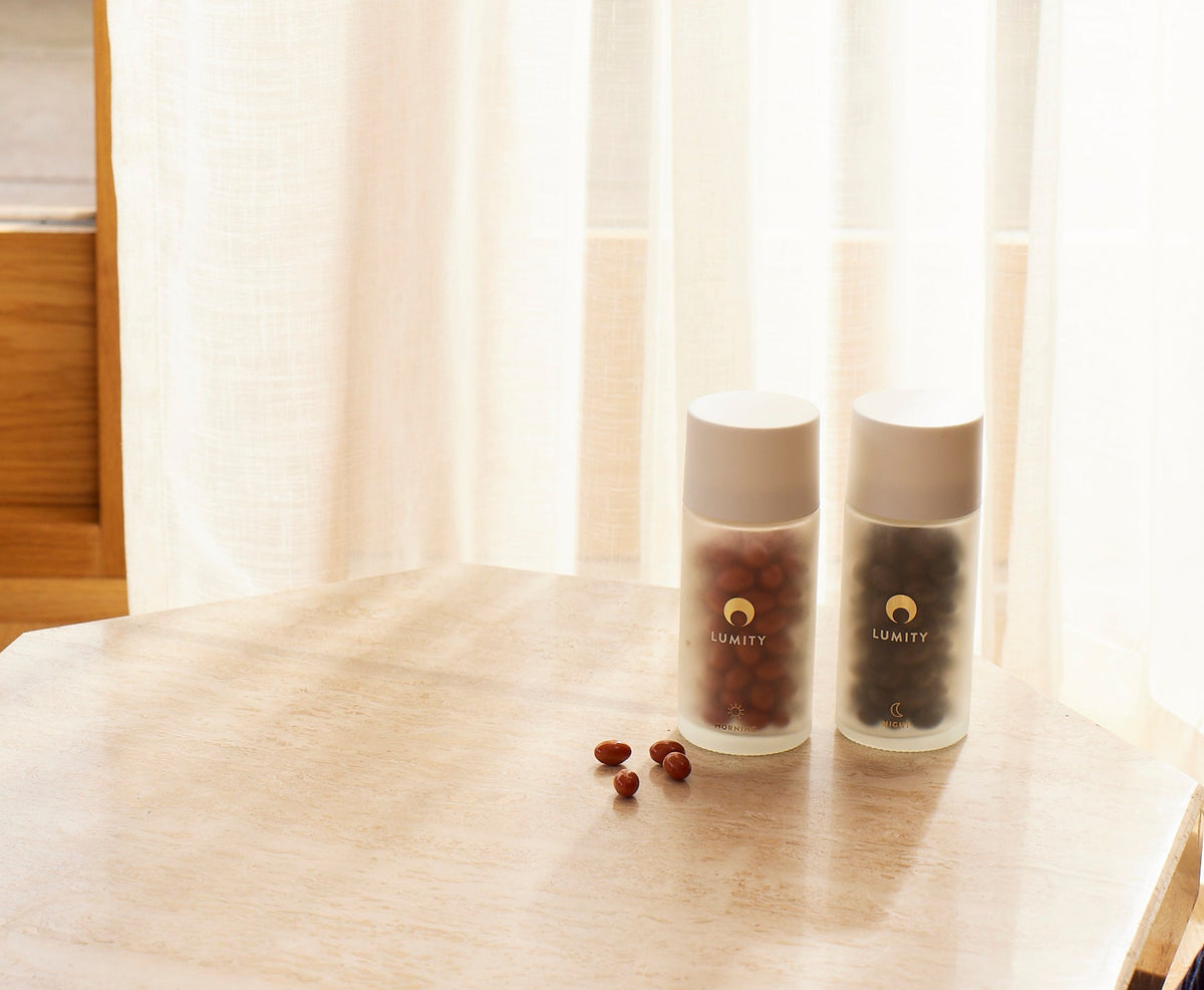 Frosted LUMITY morning & night bottles with sun/moon labels, reddish-brown morning capsules spilled beside dark night ones