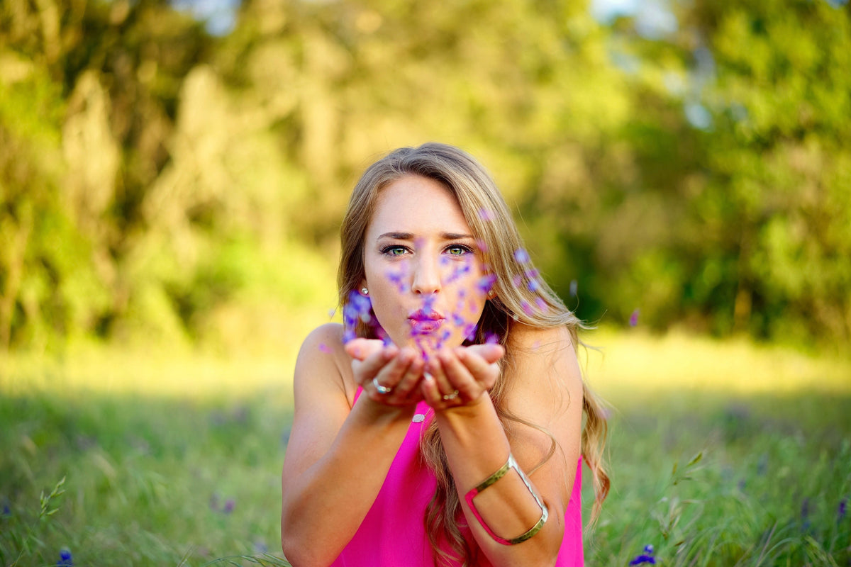 Woman in bright pink top at golden hour, blowing purple flower petals from cupped hands, metallic bracelet on right wrist.