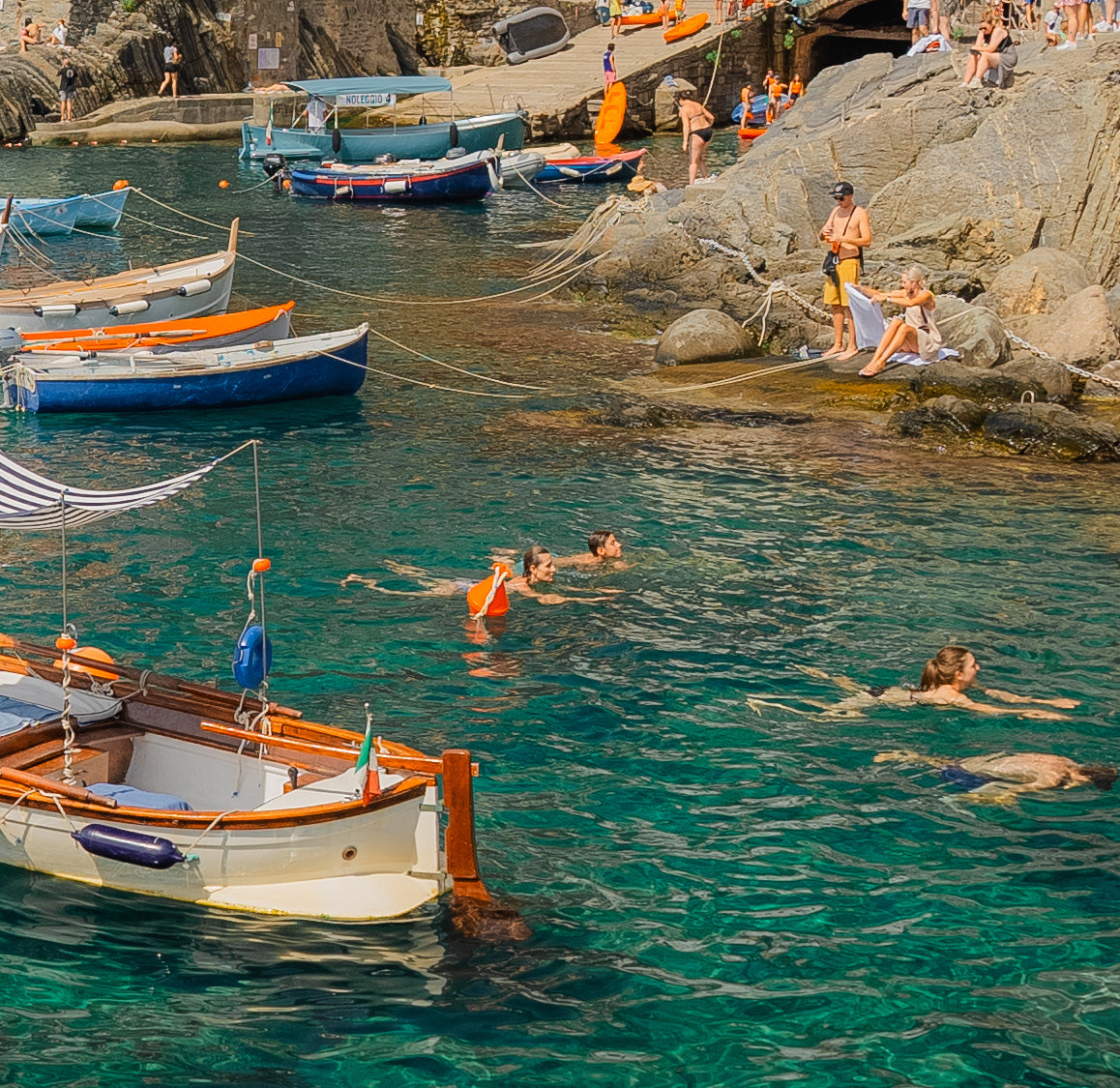 Rocky waterfront with swimmers in turquoise water, some using orange flotation devices, boats moored, people on the rocks.
